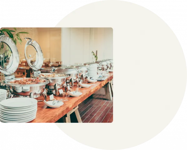 A white circle is displayed on a grey background with silver buffet trays lined across a table on the left-hand side.