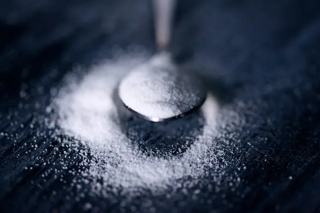 A spoon full of sugar sitting on a black table top with some sugar sprinkled around it